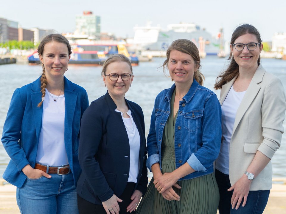 mobiliteam by NAH.SH: vier Personen stehen draußen an der Kieler Förde, das Wasser ist im Hintergrund zu sehen.