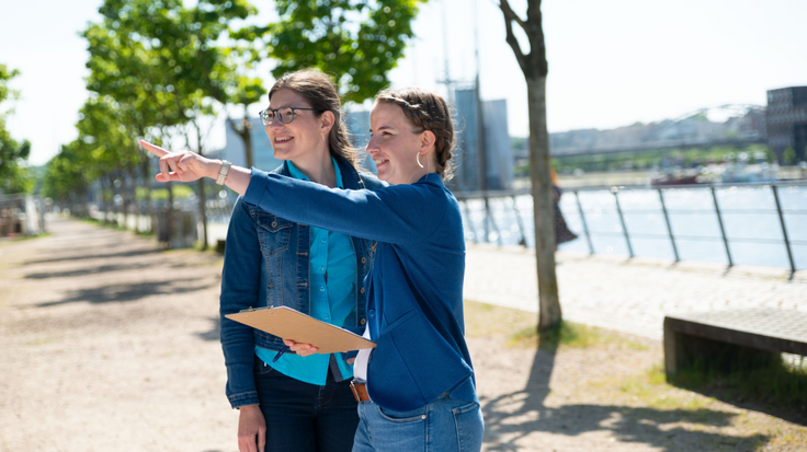 Zwei Frauen stehen bei sonnigem Wetter im Freien. Eine Frau deutet auf etwas in der Ferne.