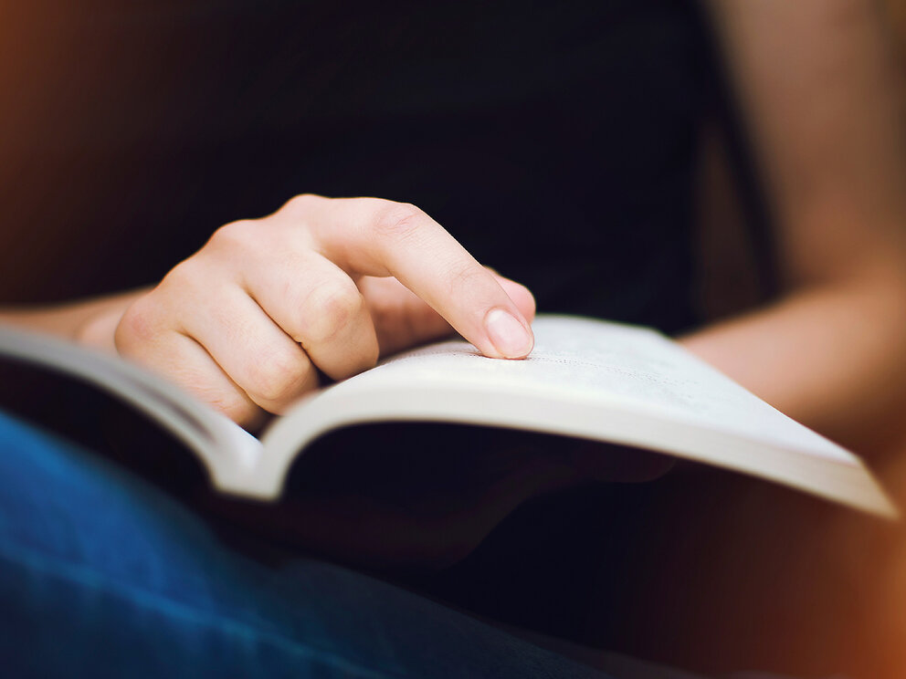 Detailaufnahme eines aufgeschlagenen Buches mit einer Hand, bei der der Zeigefinger auf einer Seite etwas markiert